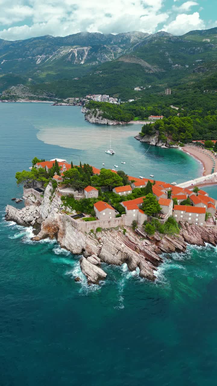 Island of Sveti Stefan - Montenegro. Aerial view. Drone going backwards. Waves breaking on the rock. Famous and luxury travel destination. Boats in the bay. Turquoise water.