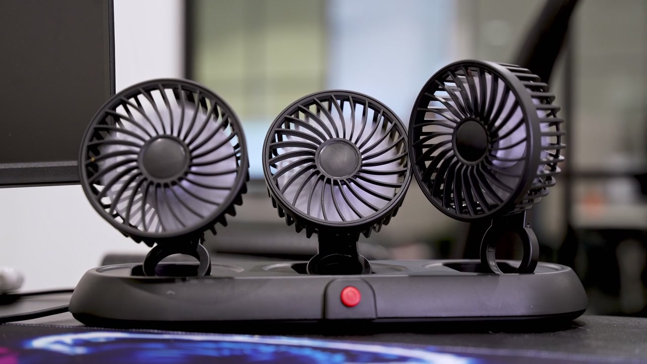 Et nærbillede af en sort mini-bordventilator med tre hoveder, der snurrer på et moderne kontorbord. Den bærbare køleenhed står ved siden af en computerskærm med en sløret indendørs baggrund.
