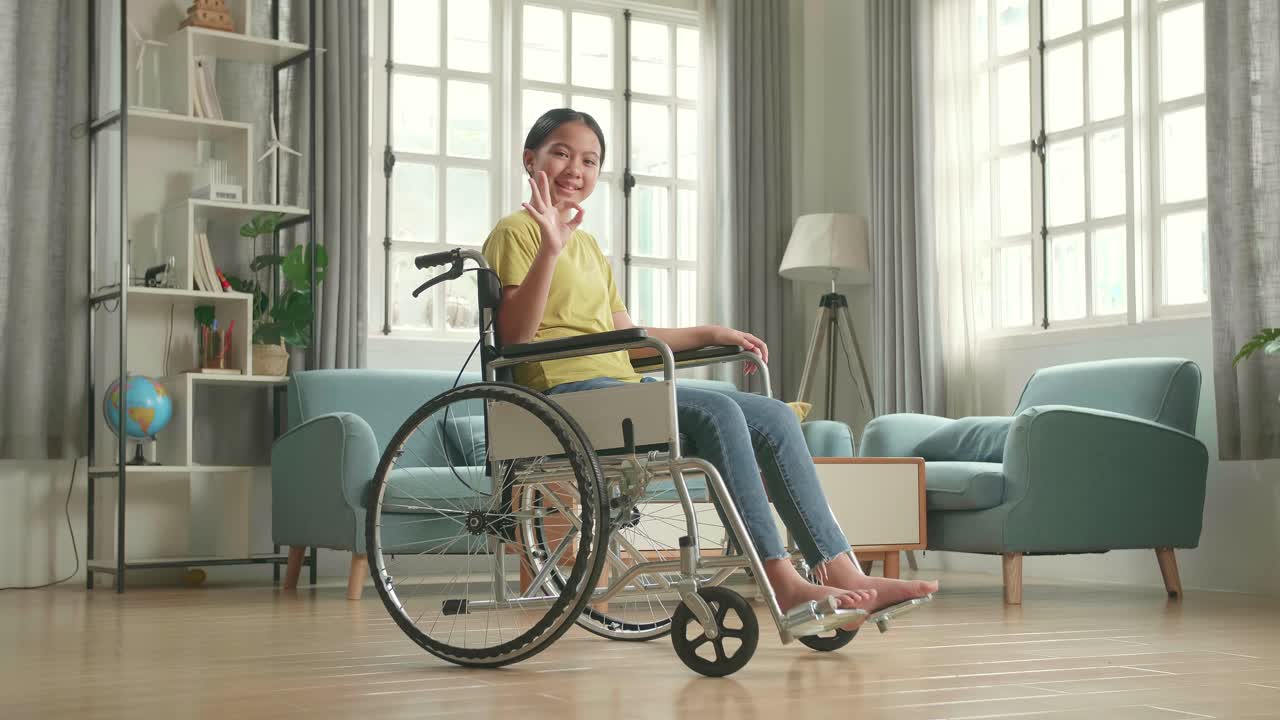 Asian Kid Girl Sitting In A Wheelchair Smiling And Showing Ok Sign In Living Room