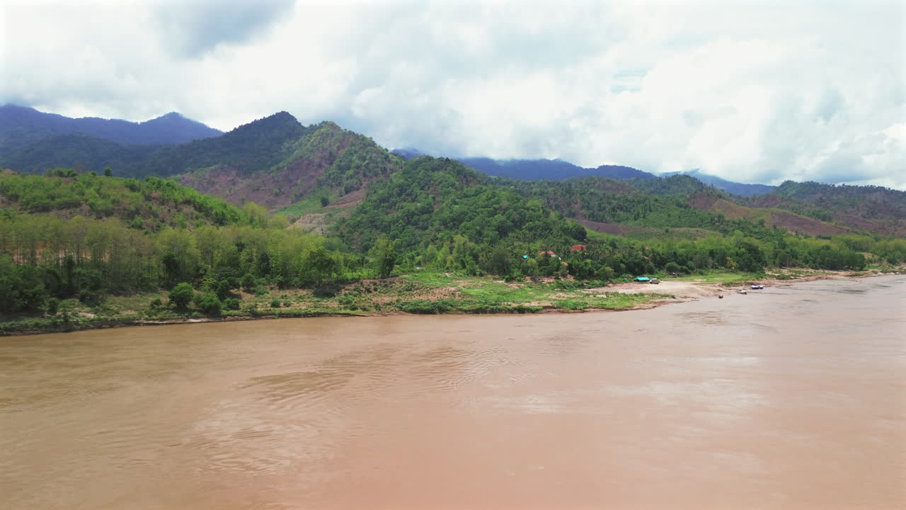 Long wooden slow boat travels along muddy Mekong River surrounded by lush green hills, aerial dolly to water