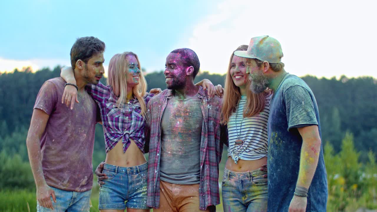 retrato del feliz y alegre equipo multiétnico de amigos en las coloridas pinturas de pie juntos en abrazos y riendo a la cámara afuera en el festival holi