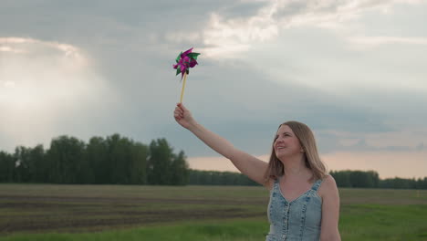 Young woman holds colourful pinwheel high while walking along grassy field edge by asphalt road under overcast summer sky hair drifting in gentle breeze conveying playful sense of freedom