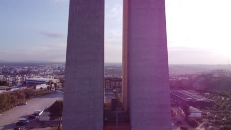 Drone-Ascendiendo-Elevándose-Alto-Sobre-La-Cabeza-De-Jesucristo-La-Base-De-La-Poderosa-Estatua-De-Cristo-Rei-En-Portugal-Lisboa-Con-La-Ciudad-Y-El-Sol-En-La-Espalda