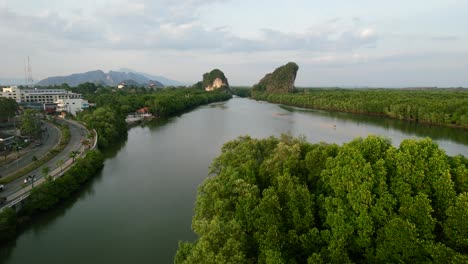 aerial drone flying low over an island forest surrounded by a river and limestone mountains as cars pass on a highway road during golden hour sunset in krabi town thailand