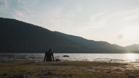 woman turns head around to relax by lake at sunset. lady tries to feel relaxation on picnic with view of mountains after hard week. calm weekend alone