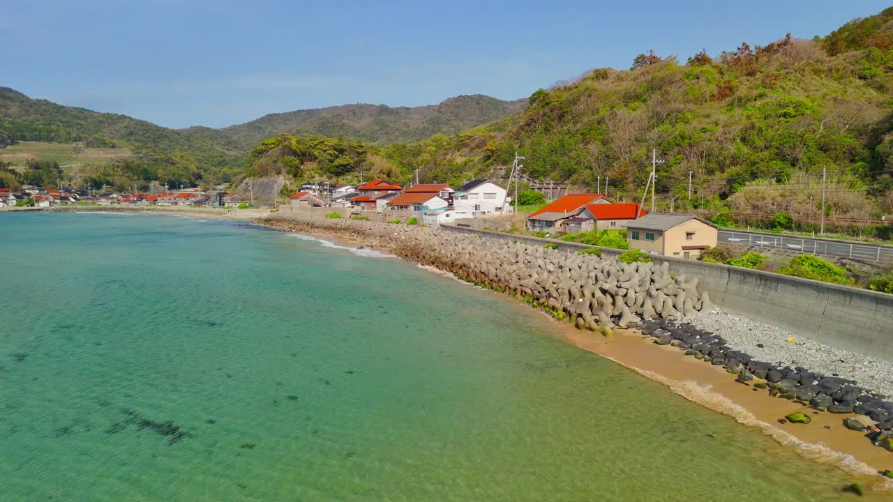 A low aerial fly-along shot captures the serene beauty of a Japanese coastal village, gliding over clear turquoise water towards the shore with lush green hills in the background