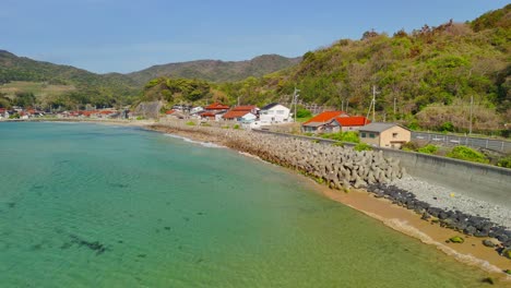 a low aerial fly-along shot captures the serene beauty of a japanese coastal village, gliding over clear turquoise water towards the shore with lush green hills in the background