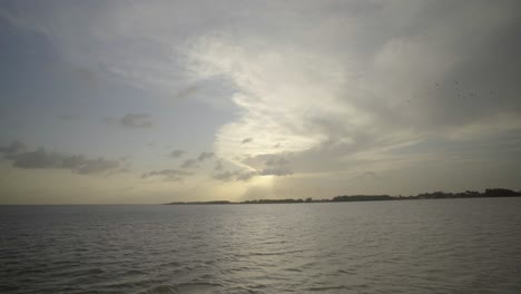 Wide-low-angle-view-of-brown,-calm,-slow-flowing-plain-river-water,-view-from-boat-under-cloudy-sky-with-landscape-silhouettes