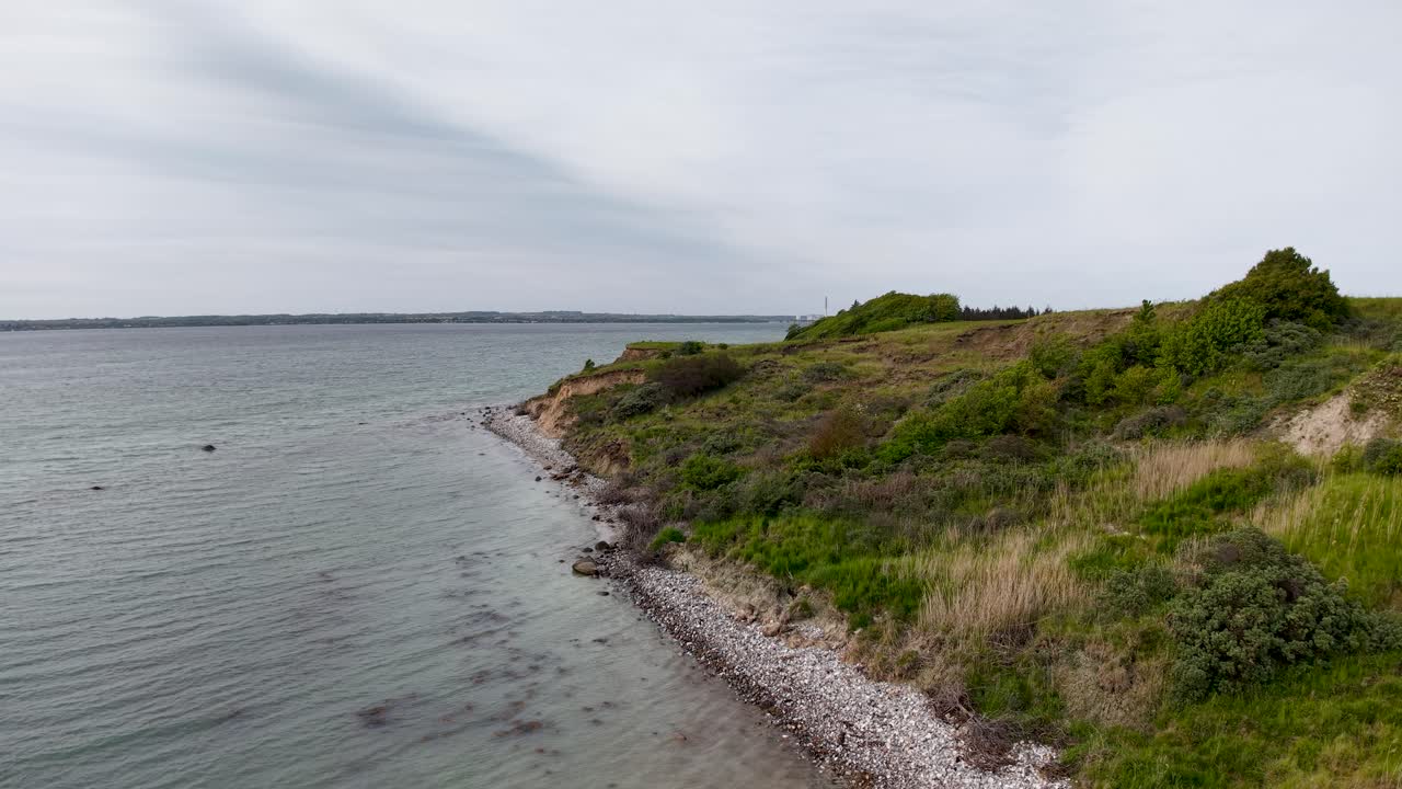 Low-altitude aerial view of a narrow coastal bluff lined with shrubs and trees, sloping into clear, shallow waters. The coastline extends into the distance under a cloudy sky