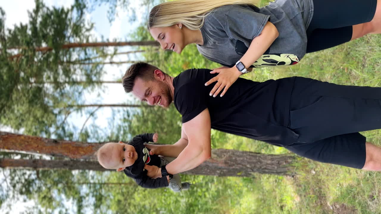 Happy Caucasian bearded man holding high his lovely baby. Smiling blonde woman stands behind looking at her son. Forest at backdrop. Vertical video.