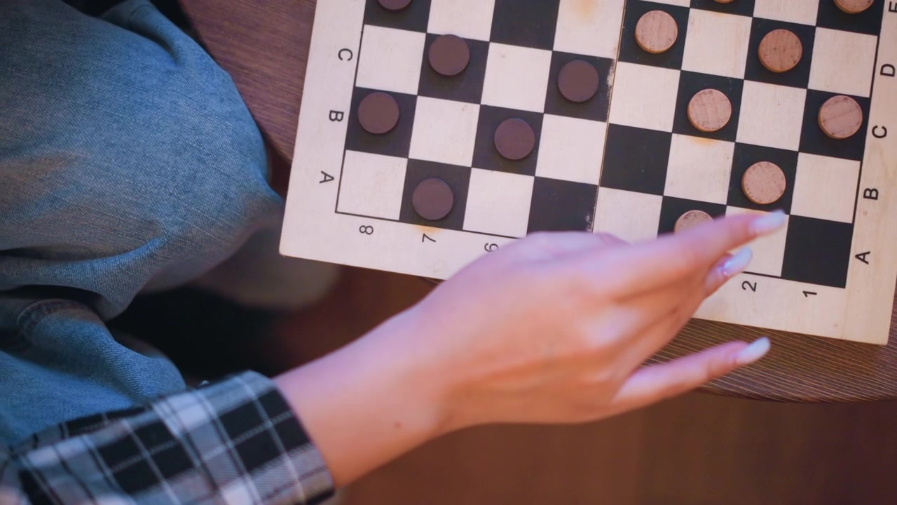 Close-up da mão de uma jovem movendo peças de jogo em um tabuleiro de damas. Mesa de madeira, jeans azul e camisa xadrez visíveis ao fundo. A iluminação suave cria tons quentes em toda a cena