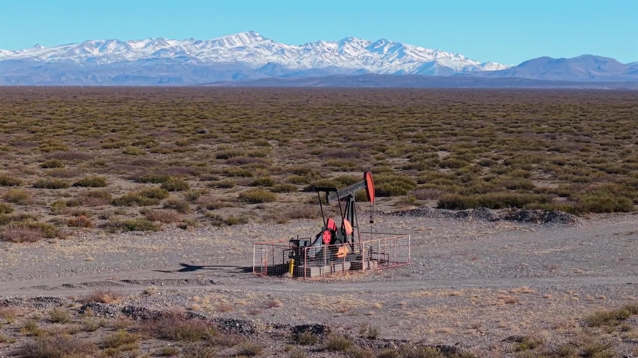 Un bilanciere di estrazione petrolifera opera a Malargüe, Mendoza, Argentina, estraendo idrocarburi da un campo di produzione remoto circondato da steppa arbustiva e Ande innevate, con drone che avanza e inclina la telecamera verso l'alto.