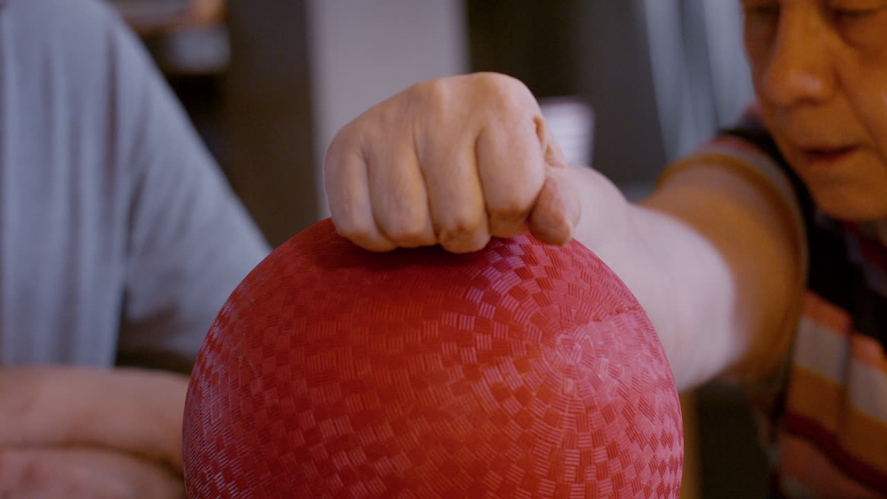 Middle-aged male caregiver gently assists the hemiplegic right hand ball therapy of an elderly female stroke survivor, seated by glass top kitchen table at home. Back, and forth hand motion 24fps.