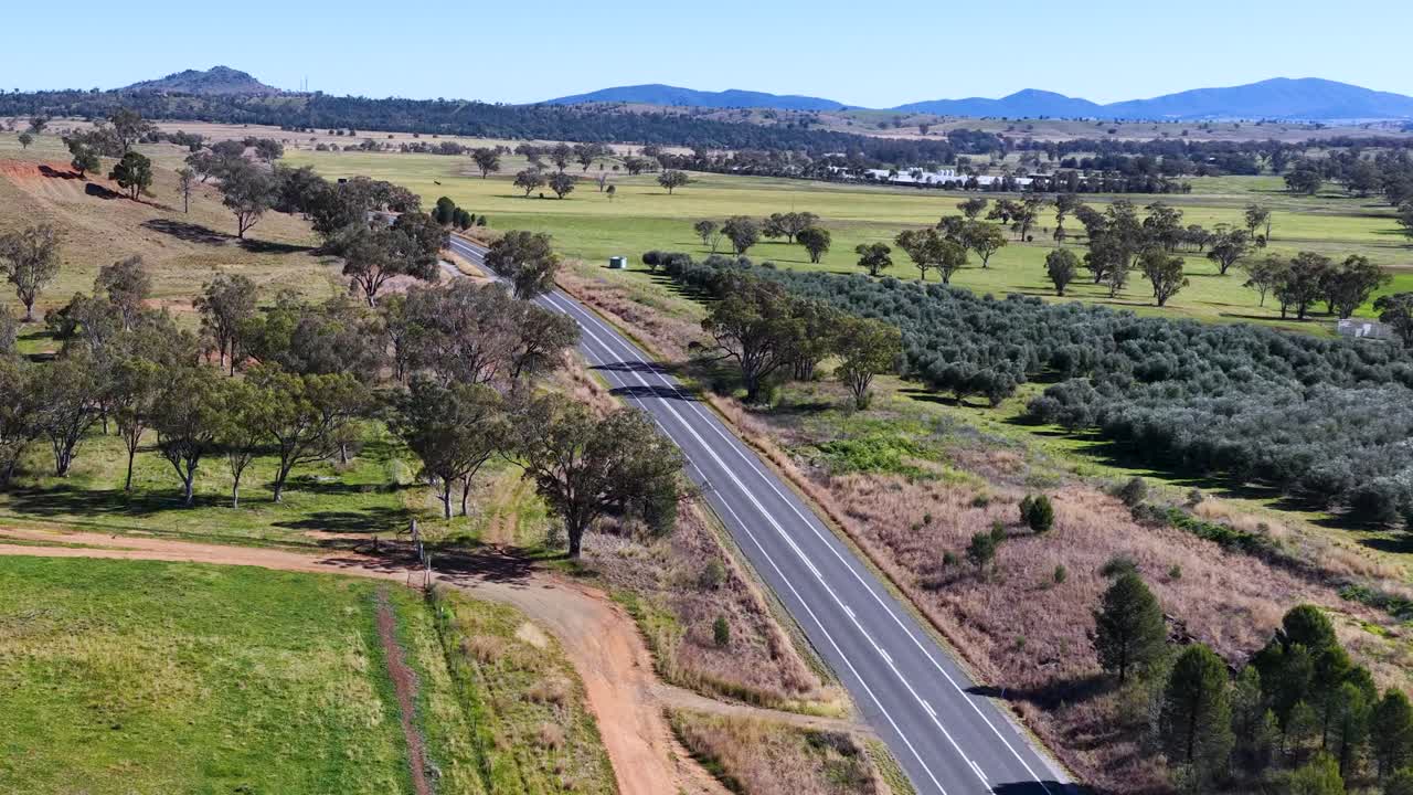 Drone footage tracks a car driving along a winding rural road bordered by fields, scattered trees, and distant hills under bright daylight