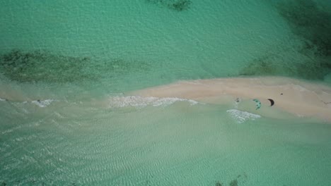 A-kite-surfer-gliding-over-turquoise-waters-near-a-sandy-beach,-aerial-view