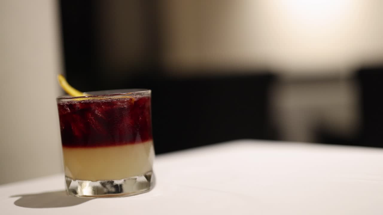 Uma mão alcança um coquetel vibrante sobre uma mesa, destacando um ambiente relaxante e elegante com iluminação suave.