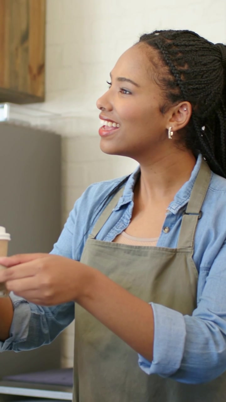 Vertical video: Laughing barista handing paper cup at counter after customer call in olive apron