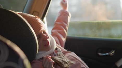 a young girl in a pink knit hat and scarf is stretching her hands and body while sitting in a car seat. the scene captures a moment of awakening and comfort during a car ride on a cold day