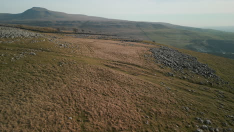 Flying-over-rocky-hillside-revealing-green-valley-with-mountain-Ingleborough-on-horizon-at-Ingleton-Yorkshire-UK