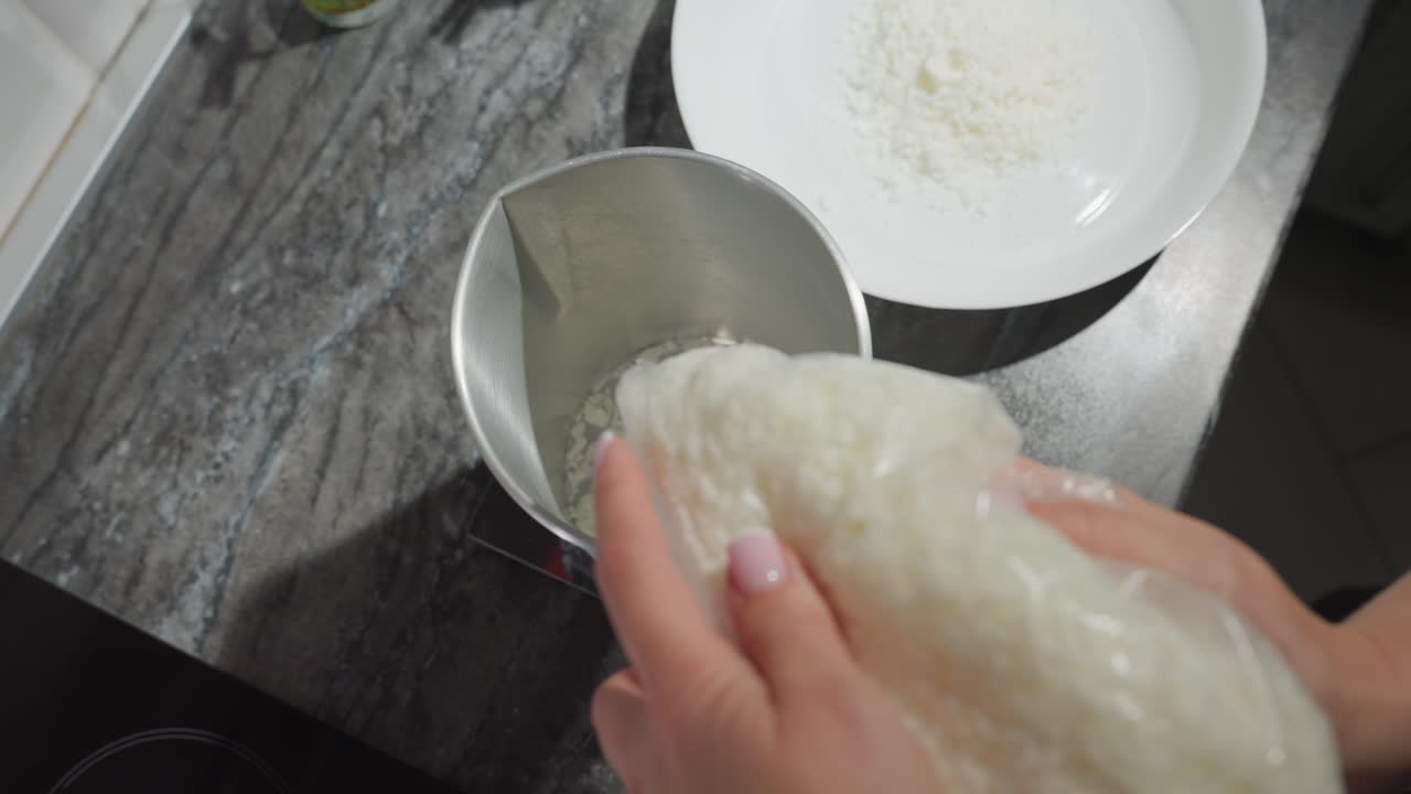 Female chef hand pouring sugar from transparent bag into metal jar on food scale beside white plastic plate with flour on countertop in kitchen preparing ingredients for baking