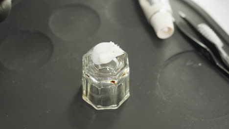 close-up view of technician squeezing cream into small transparent glass jar on black silicone tray with beauty tools nearby, preparing to mix cosmetic substance for treatment session