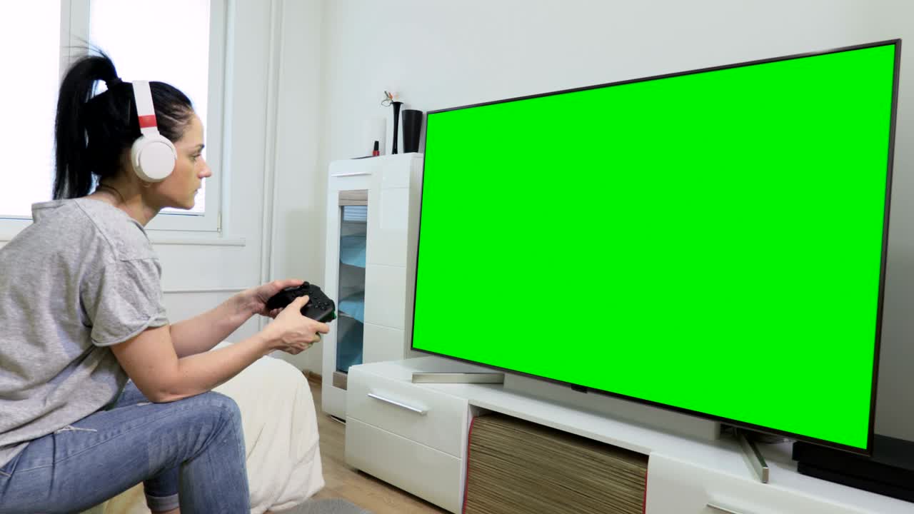 녹색 화면 근처에서 비디오 게임을 하기 위해 헤드폰과 게임 콘솔을 사용하는 여성