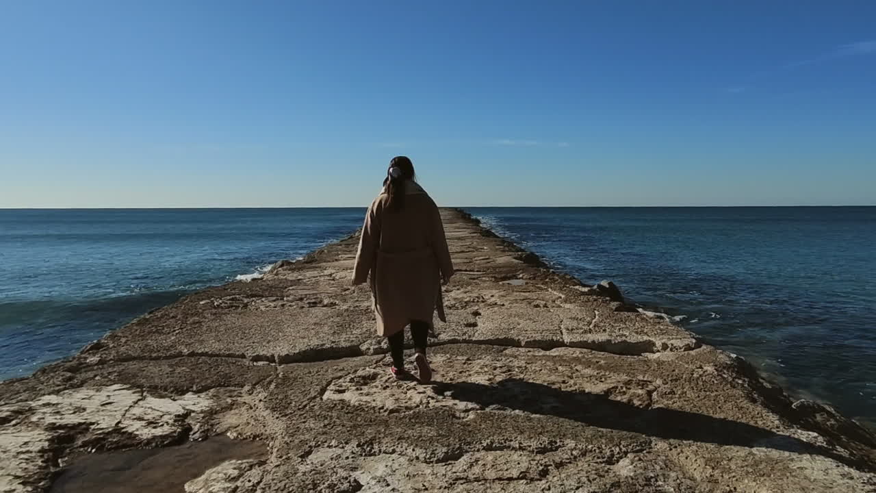 Tracking rear shot of woman in coat walking on rocky jetty towards the ocean during sunny day with blue sky