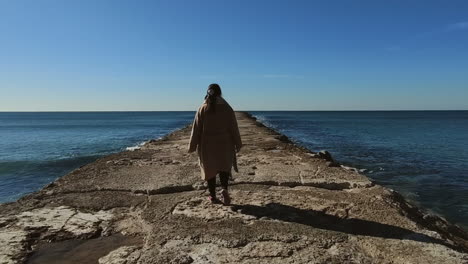 Seguimiento-De-La-Toma-Trasera-De-Una-Mujer-Con-Abrigo-Caminando-Sobre-Un-Embarcadero-Rocoso-Hacia-El-Océano-Durante-Un-Día-Soleado-Con-Cielo-Azul