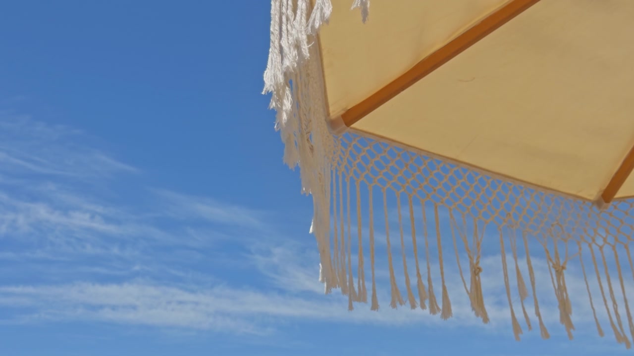 Splendida ripresa 4K dal basso di ombrelloni in macramè bianchi con frange in un resort di lusso sulla spiaggia. Presenta una fila organizzata di lettini, palme tropicali e un cielo azzurro limpido
