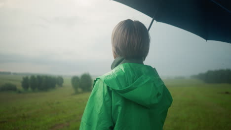 back view of woman in green raincoat holding an umbrella while standing in a vast grassy field, looking into the distance, the sky is cloudy, and trees can be seen far in the background