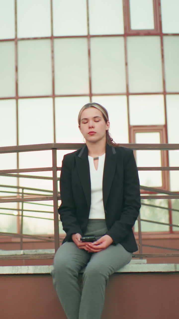 Professionista in blazer nero seduta su una recinzione con un telefono in mano, edificio moderno sullo sfondo con design urbano, illuminazione naturale, gambe incrociate alla caviglia