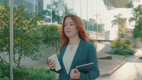 close up shot of a young caucasian woman going to office with a tablet device in hands and drinking coffee while walking along the sidewalk on a sunny morning