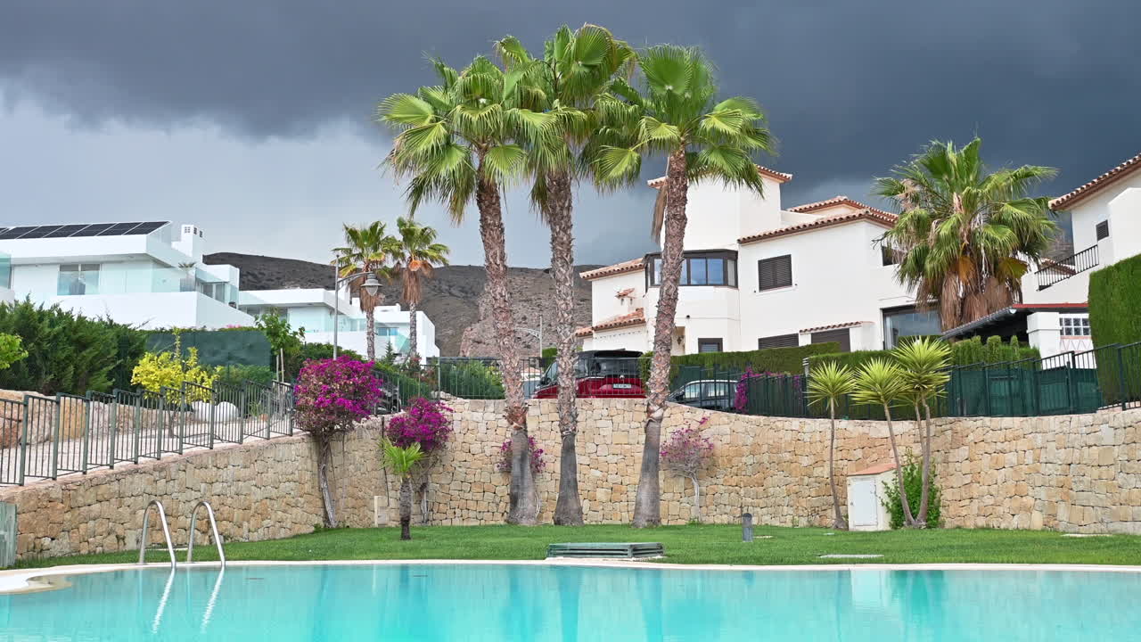 Houses and palm trees near water pool in Spain with black skyes before rain