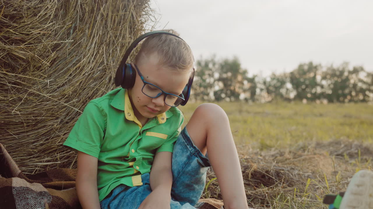 молодой мальчик с наушниками наслаждается музыкой, сидя на одеяле на открытом воздухе, спокойная сельская обстановка с сеном на заднем плане, мирный момент в сельской местности