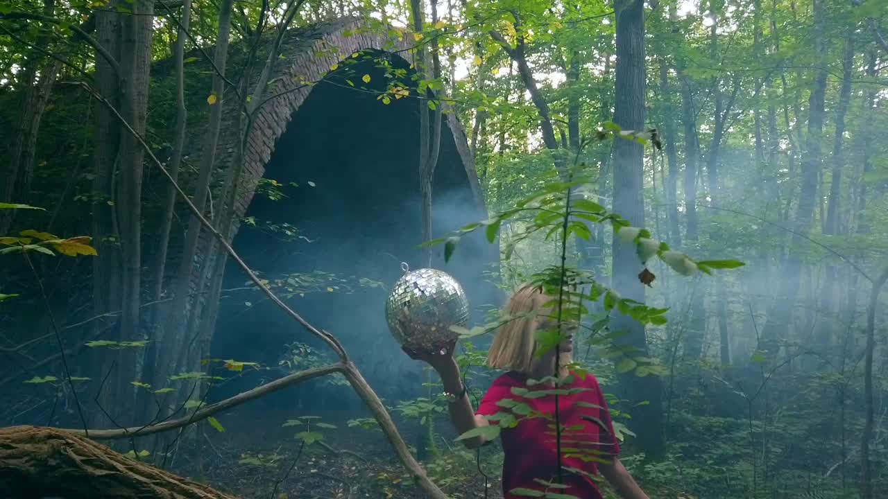 Wonderful slow motion cinematic shot of in foggy forest sitting blond jung girl in a red dress holding a mirror ball in one hand. Foto video shoot on lost places in nature 1080, 24p by Philipp Marnitz