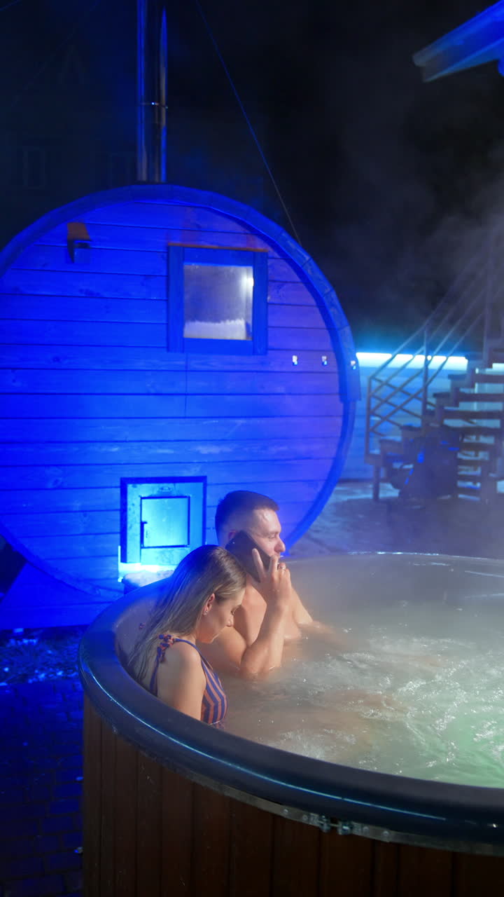 Man and woman have a spa evening in the hot tub together. Bearded man is speaking on the phone sitting in jacuzzi. Vertical video.