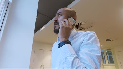 hombre indio asiático caminando hacia la ventana de la oficina hablando por teléfono móvil