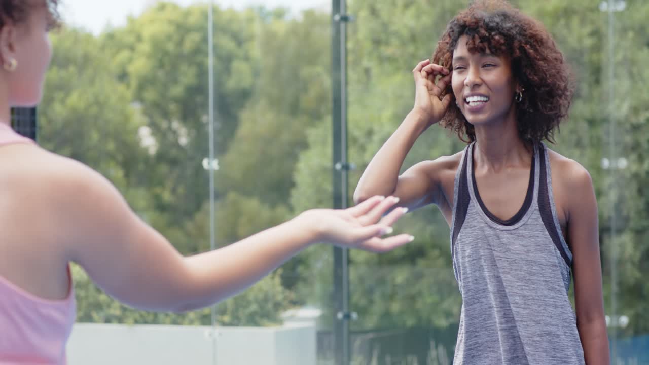 Mujeres afroamericanas estrechándose las manos en una cancha de cristal, la mujer con camiseta de tirantes gris cogiendo una pala para jugar, con espacio para texto.