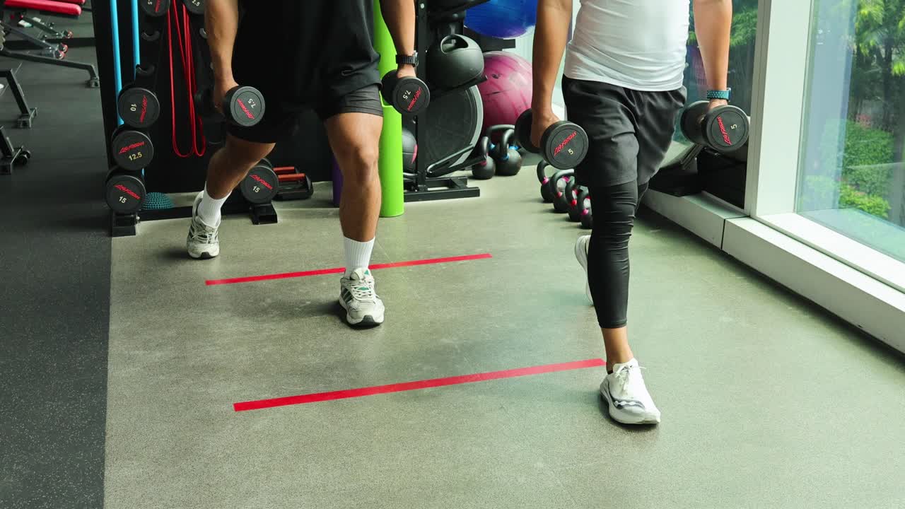 Two men perform synchronized dumbbell walking lunges in a modern sunlit gym setting