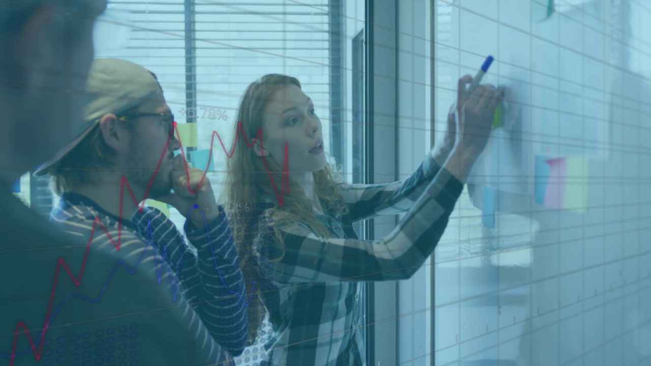 Female colleague writing on glass panel with notes in tech office, showing floating chart graphics
