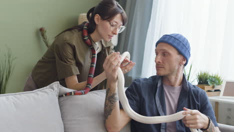 couple holding pet snakes and talking together while the man is sitting and woman is leaning on a comfortable sofa at home 2