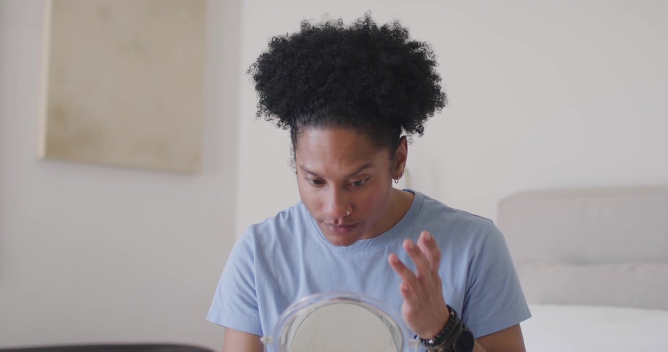 Uomo africano che nota un'imperfezione, seduto sul letto con una maglietta blu, che tiene uno specchio, prendendosi cura del viso.