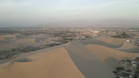 Massive-desert-sand-dunes-tower-high-above-hazy-Ice-Peru,-aerial