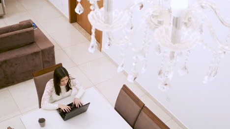 caucasian woman drinks coffee and types on laptop at homework, under chandelier