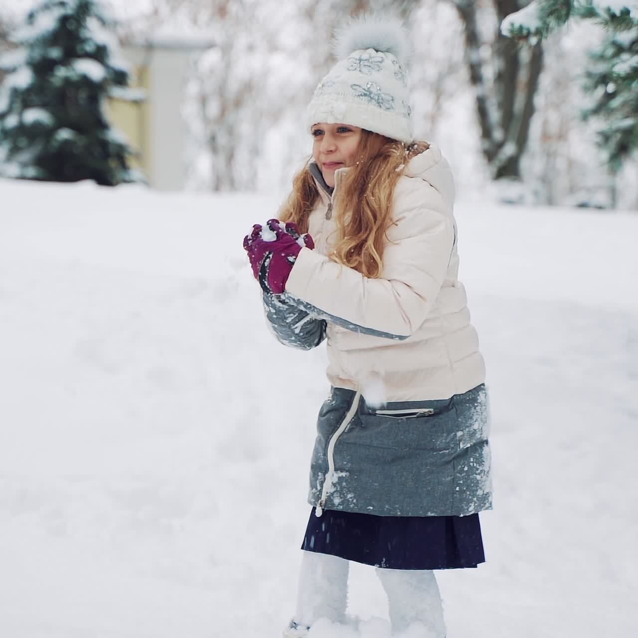 Smiling girl with long hair is throwing snowballs in the street in the yard during a snowfall in the winter. Slow motion