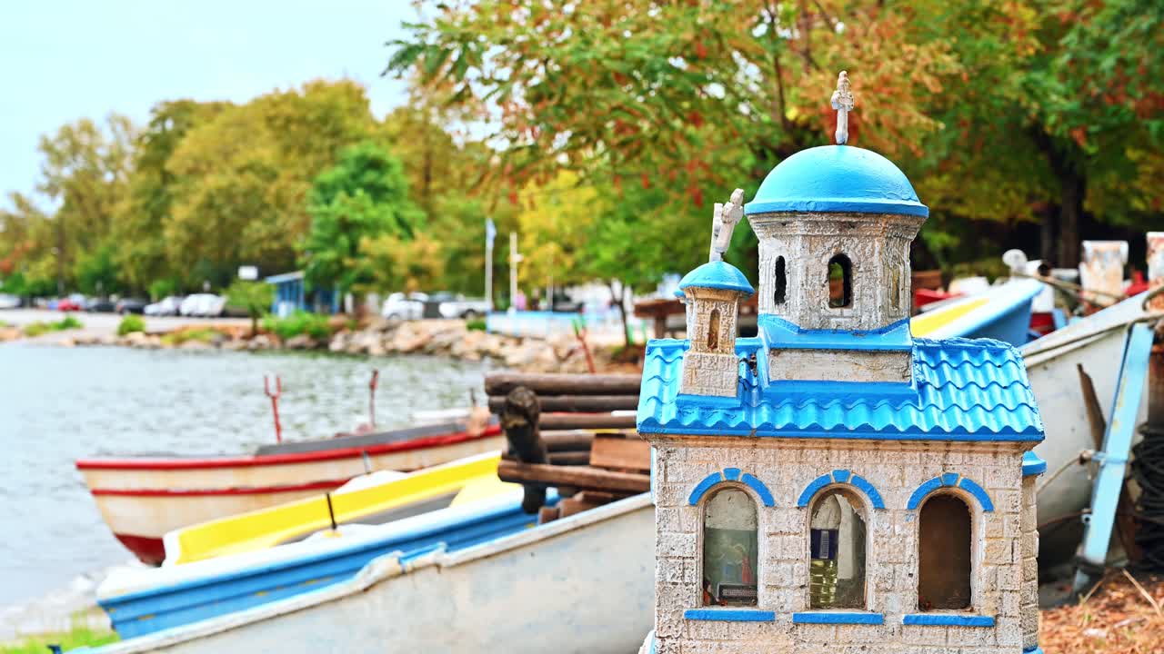 Small model of a church with blue roof and cupolas on the beach of Stavros, greenery and boats on the background, Greece