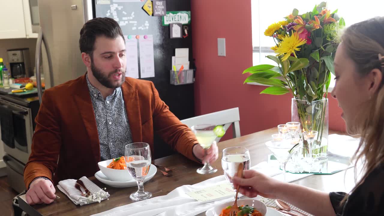 Couple Eating Orange Pasta On A Home Dining Room Table. Man Pours Cocktail Drink Into The Woman's Glass. medium shot