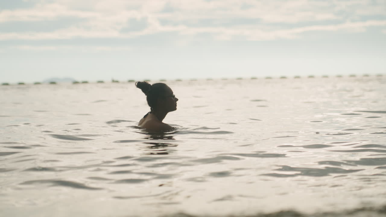 Black woman wading in shallow sea, standing silhouette against soft sunrise sky gentle tide, low waves and distant horizon create calm morning ritual vibe for wellness seeker, slow breath and quiet