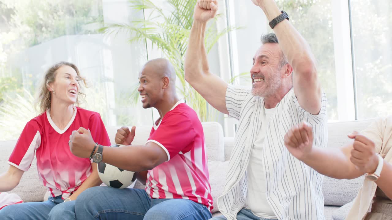 Diverse friends holding soccer ball on sofa watching at home, reacting to off-screen goal, cheering