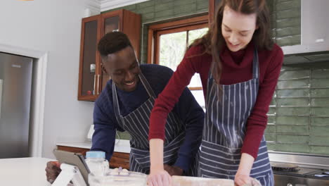 Feliz-Pareja-Diversa-Parada-En-La-Cocina,-Usando-Tableta-Y-Extendiendo-Masa,-Cámara-Lenta